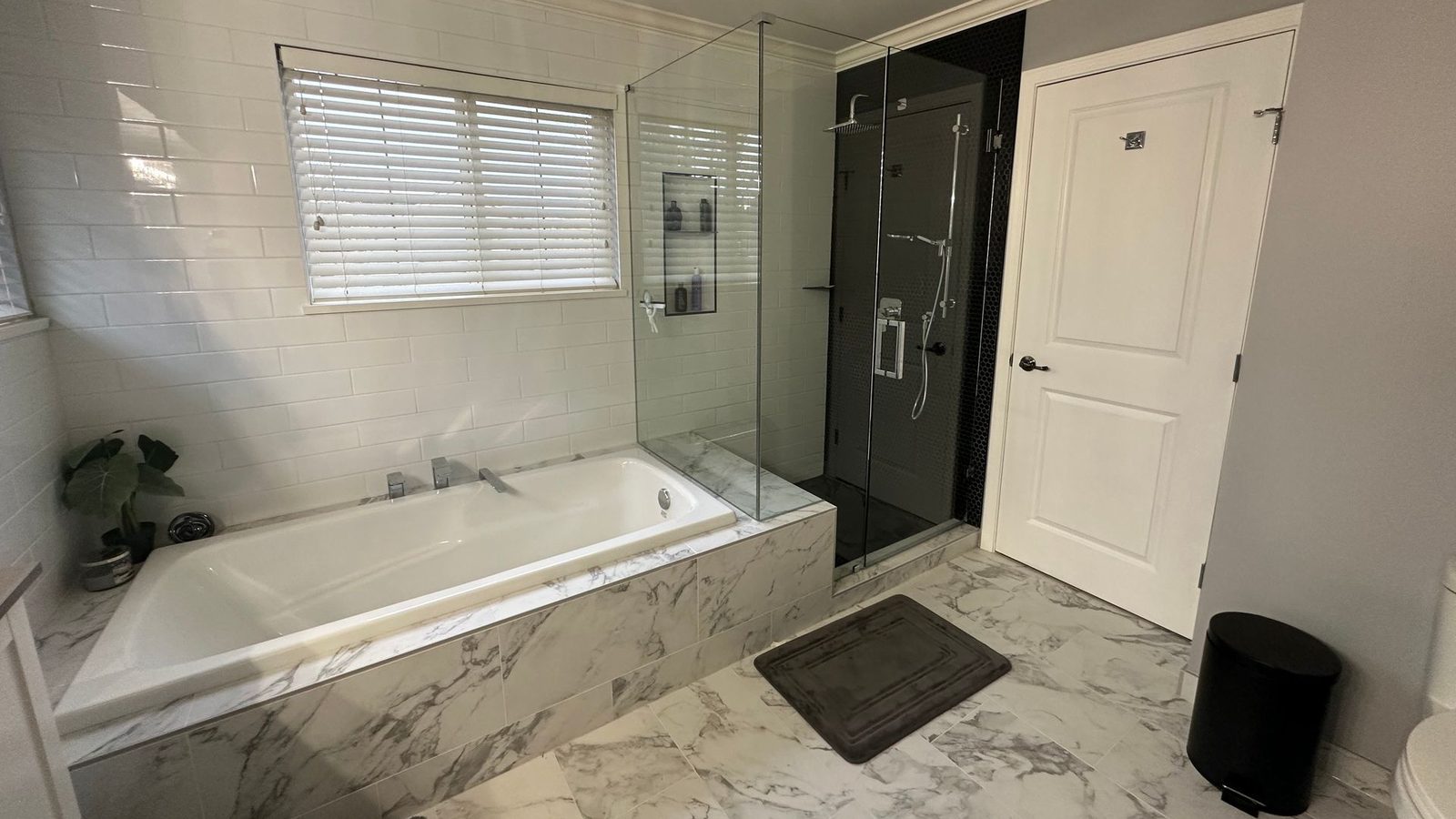 A finished bathroom with a soaker tub in a marble-look tile surround, a glass-walled shower with a black tile back wall, white subway tile on the wall and a white panelled door