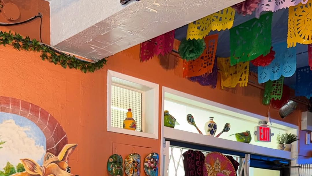 A corner of Adelicia's dining room: orange walls, strings of coloured papel picado across the ceiling, a shelf of painted Mexican pottery, three ceramic masks and a sombrero