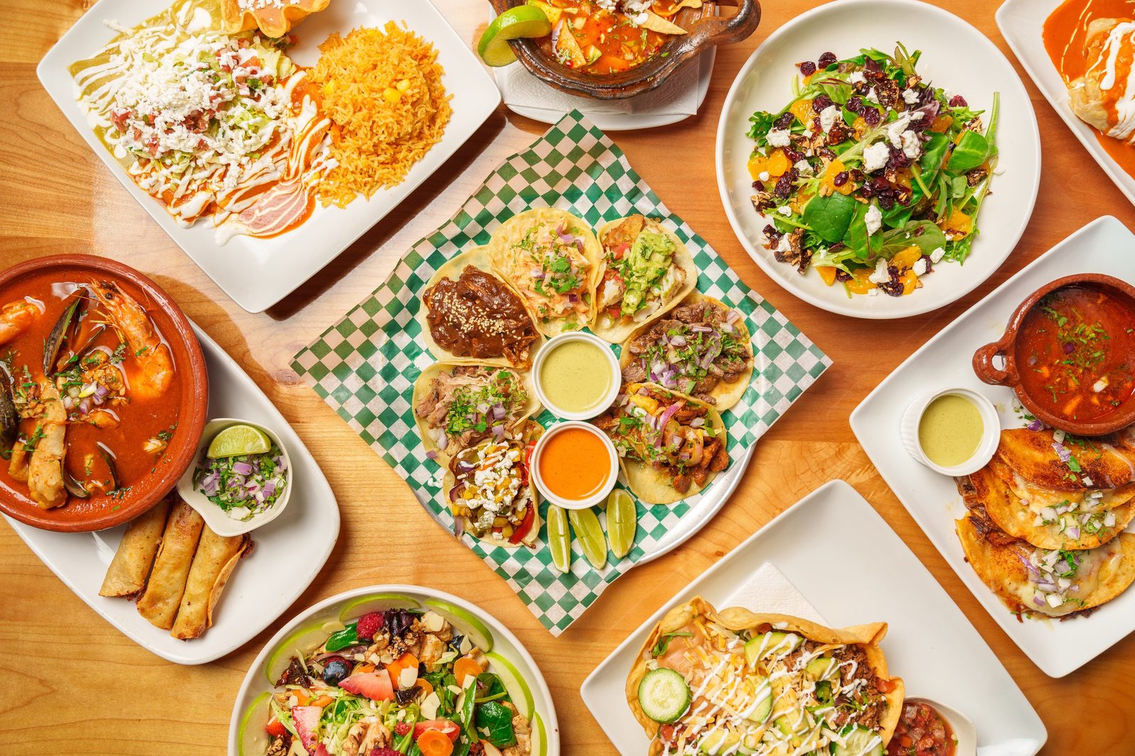 A table seen from above covered in plates: a taco platter on a green checkered paper in the middle, enchiladas with rice, a salad, a bowl of seafood soup and quesadillas