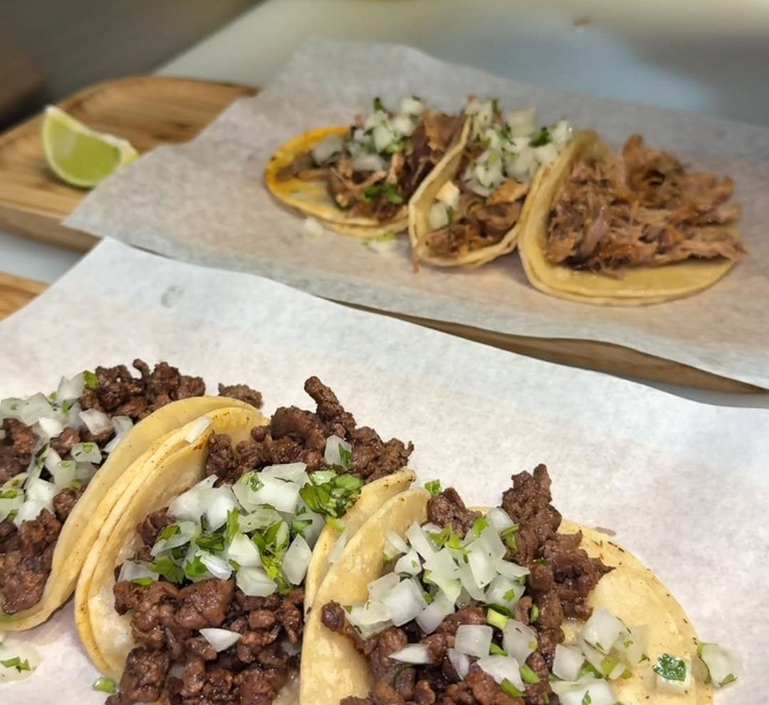 Two rows of tacos on parchment paper: pulled pork tacos at the back and grilled steak tacos in front, topped with chopped onion and cilantro, with a lime wedge