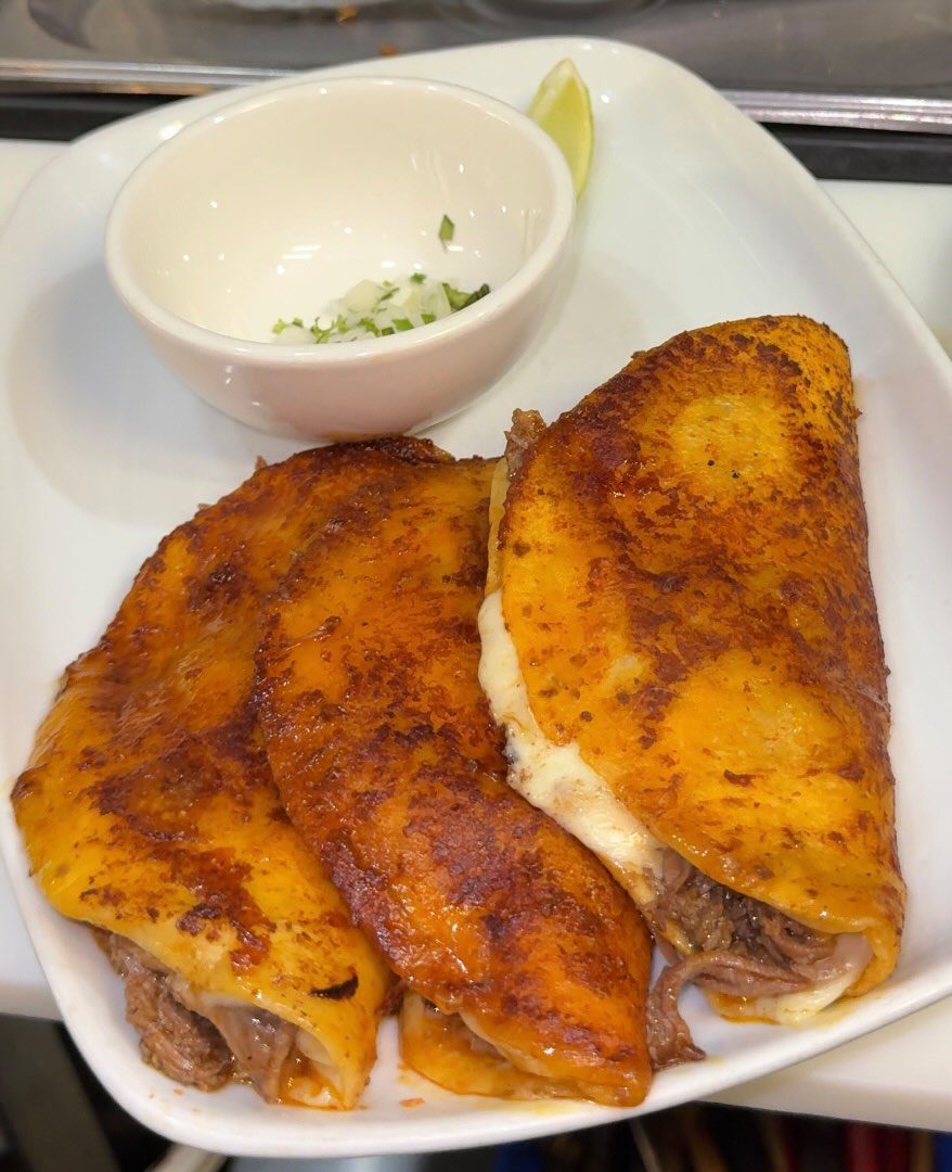 Three folded birria tacos with a crisp, orange cheese-and-chile shell on a white plate, beside a small dish of chopped onion and cilantro