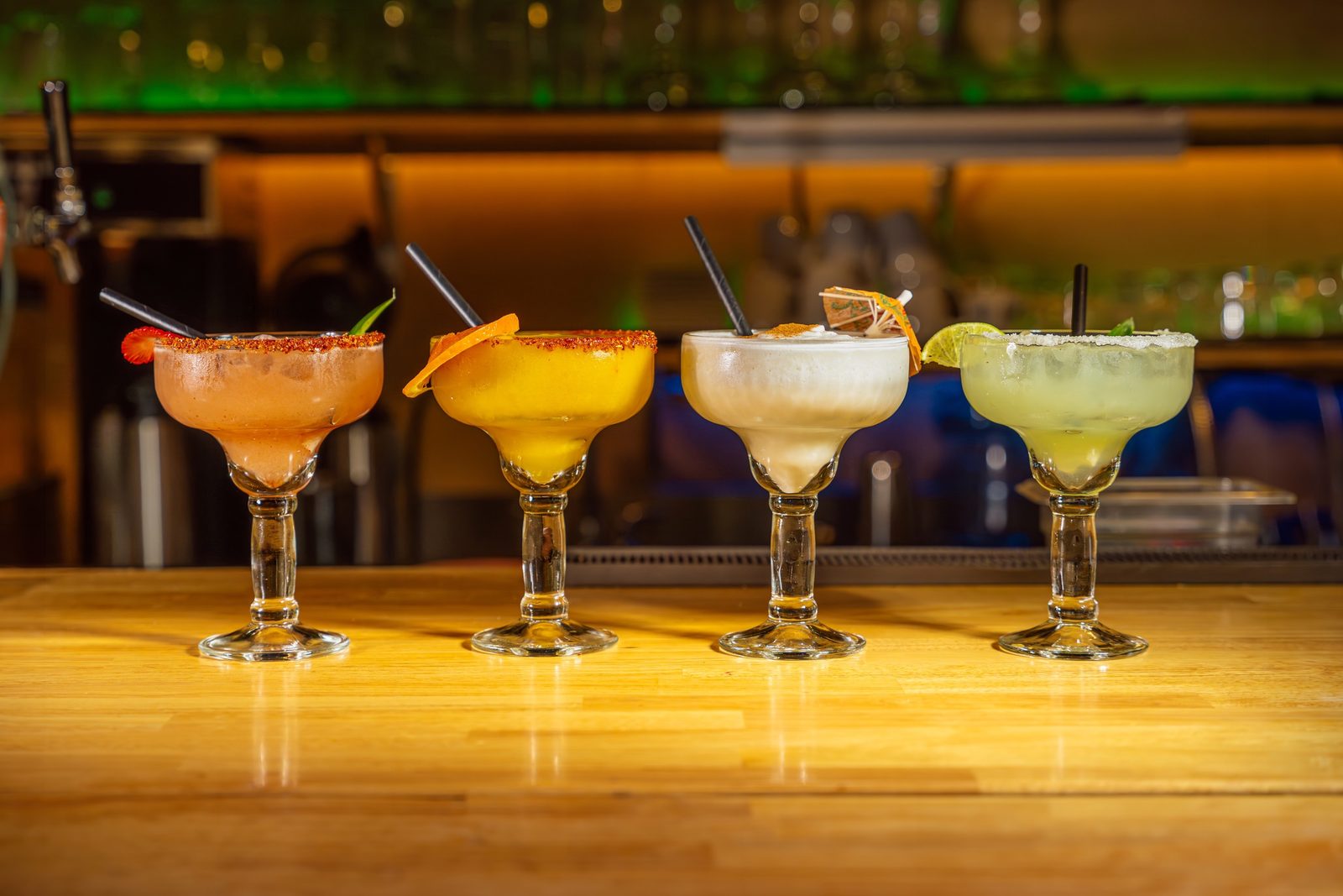 Four margaritas in different colours lined up on a wooden bar counter, each with a salted or chilli rim, with bottles glowing behind