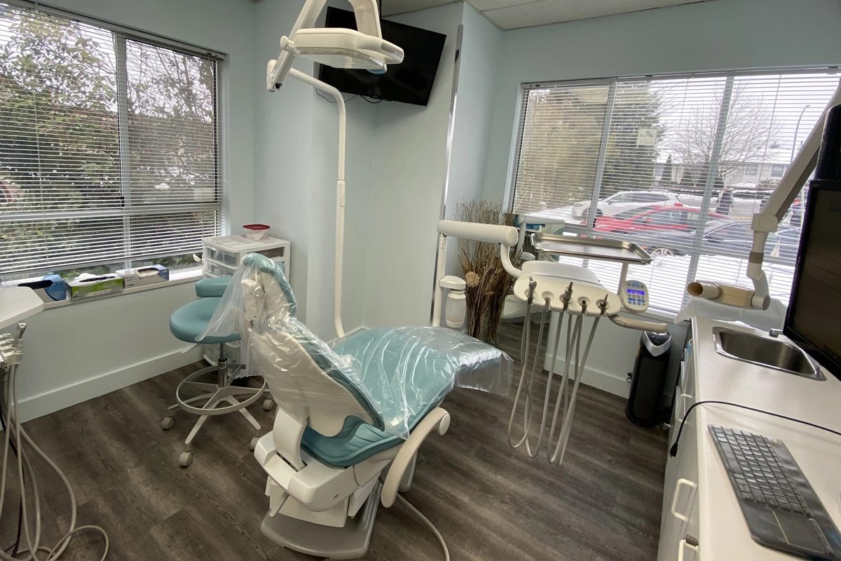 A treatment room at Allard Dental Centre with a teal dental chair, overhead light, wall-mounted screen and two large windows