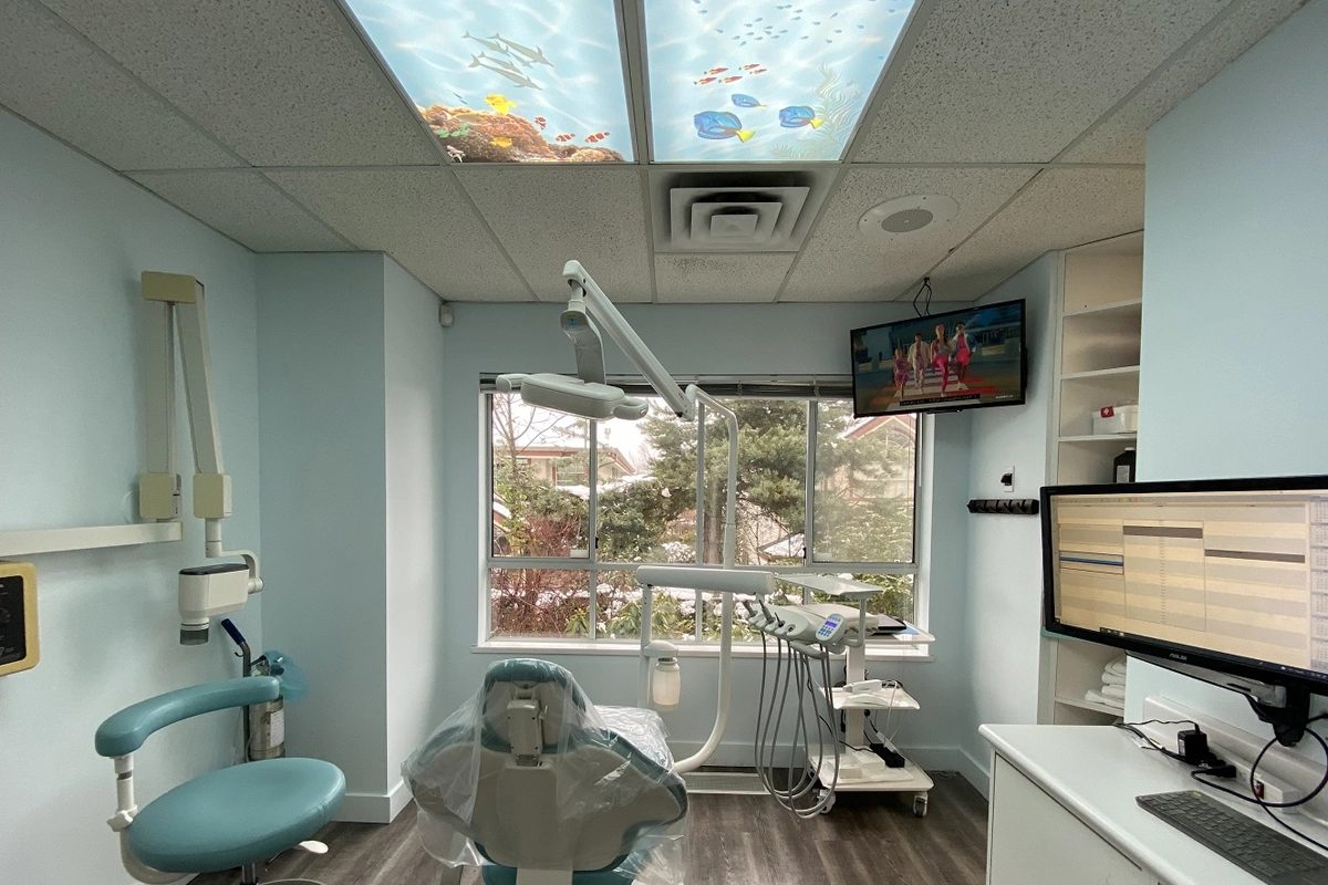 Another treatment room with ocean-scene panels in the ceiling lights, a teal chair, a computer monitor and a window onto trees