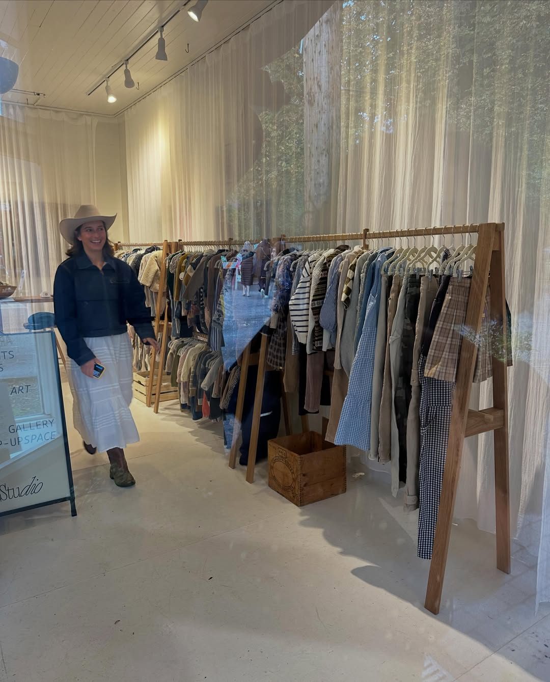 Inside Always, Sunny Shop: a long rail of consignment jeans, jackets and knits under soft white curtains, with a shopper browsing in a cowboy hat