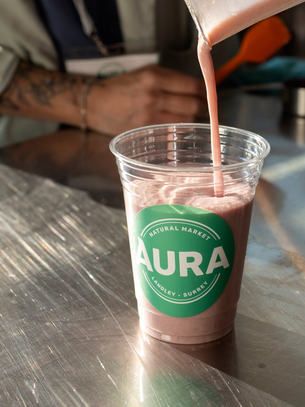 A pink berry smoothie being poured into a clear cup with a green Aura Natural Market label at the smoothie counter