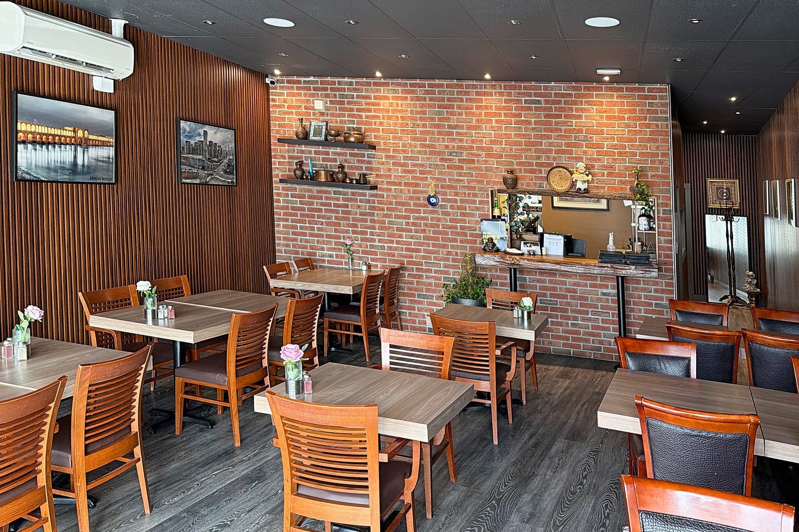 Avishan's dining room on Douglas Crescent: wood-slat walls with framed photos, a brick feature wall with shelves of copper pots, and wooden tables set with small vases of roses