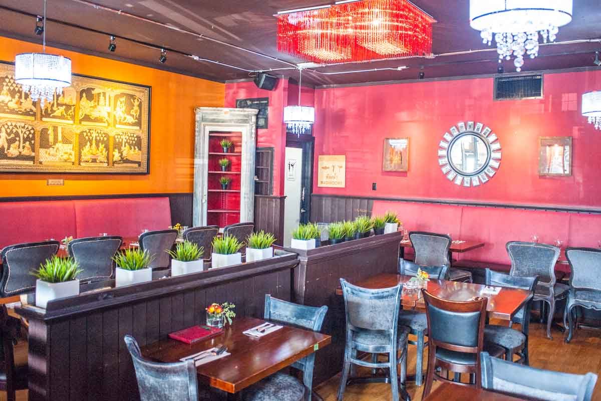 The dining room at Ban Chok Dee in Langley City: orange and raspberry-pink walls, a gold Thai temple-scene panel, a red beaded chandelier, planters on a dark wood divider and wooden tables set with napkins