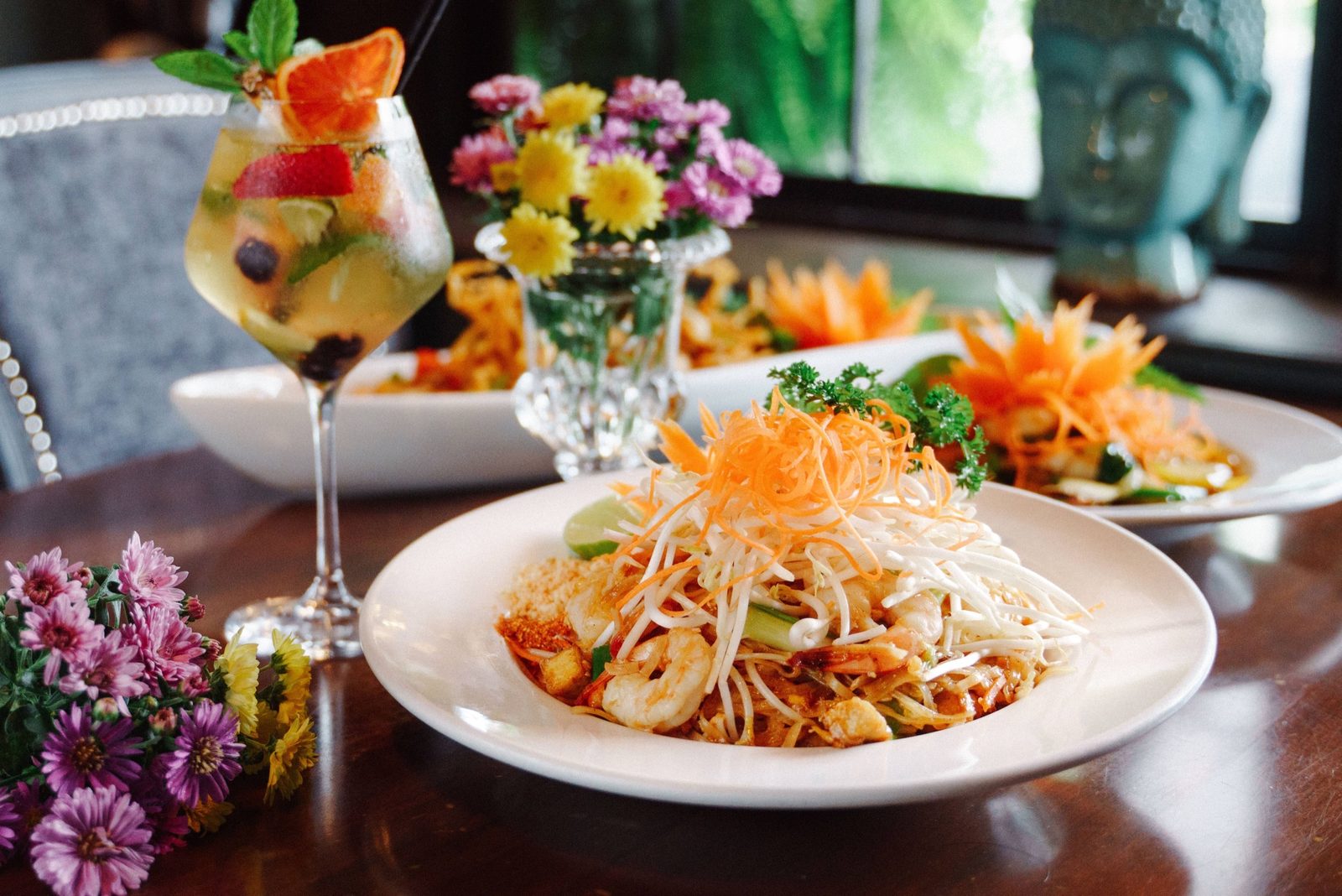 A plate of pad thai with prawns, bean sprouts and shredded carrot on a dark wood table, with a fruit-filled glass and yellow and pink flowers beside it