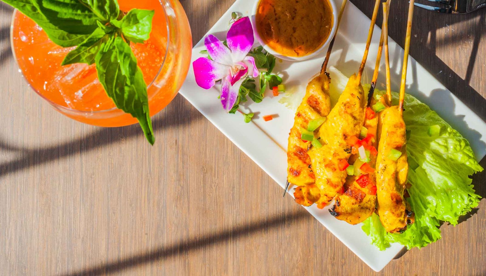 Chicken satay skewers on a white plate with peanut sauce and a pink orchid, seen from above next to a basil-topped orange drink