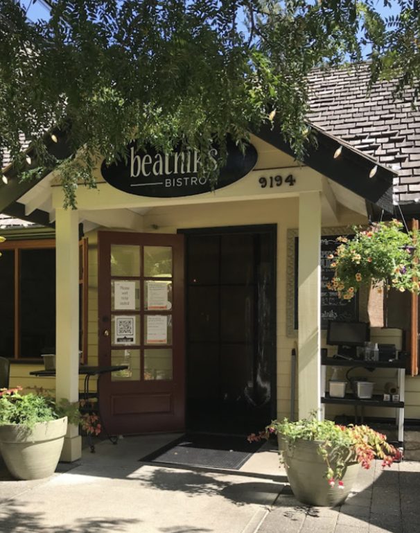 The covered front porch of Beatniks Bistro in Fort Langley: a cream house with a brown glass-panelled door, an oval sign reading beatniks bistro, the number 9194, a hanging flower basket and two stone planters