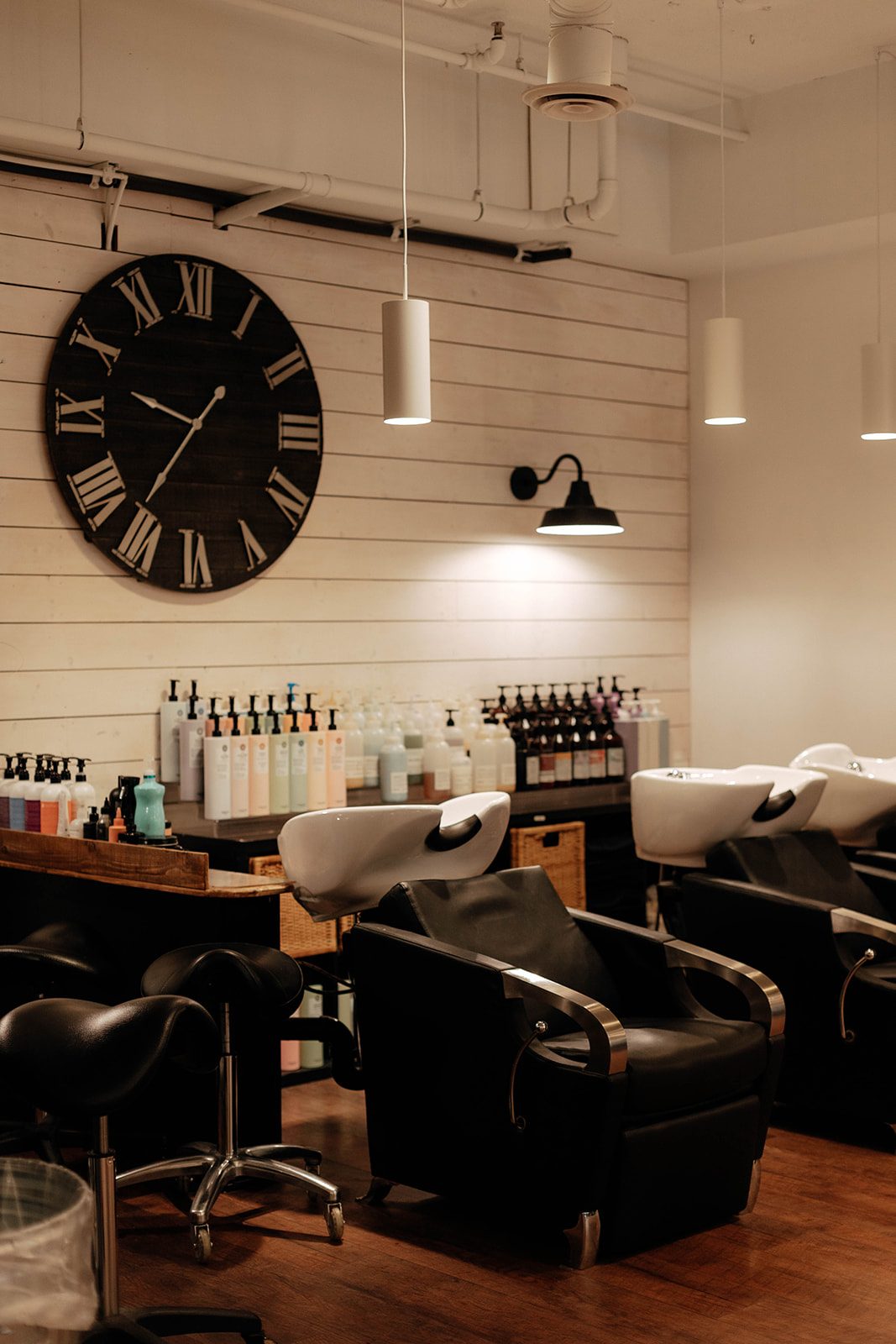 Black Orchid Hair Studio's shampoo area: a large Roman-numeral clock on a white plank wall above rows of salon bottles, white basins and black chairs