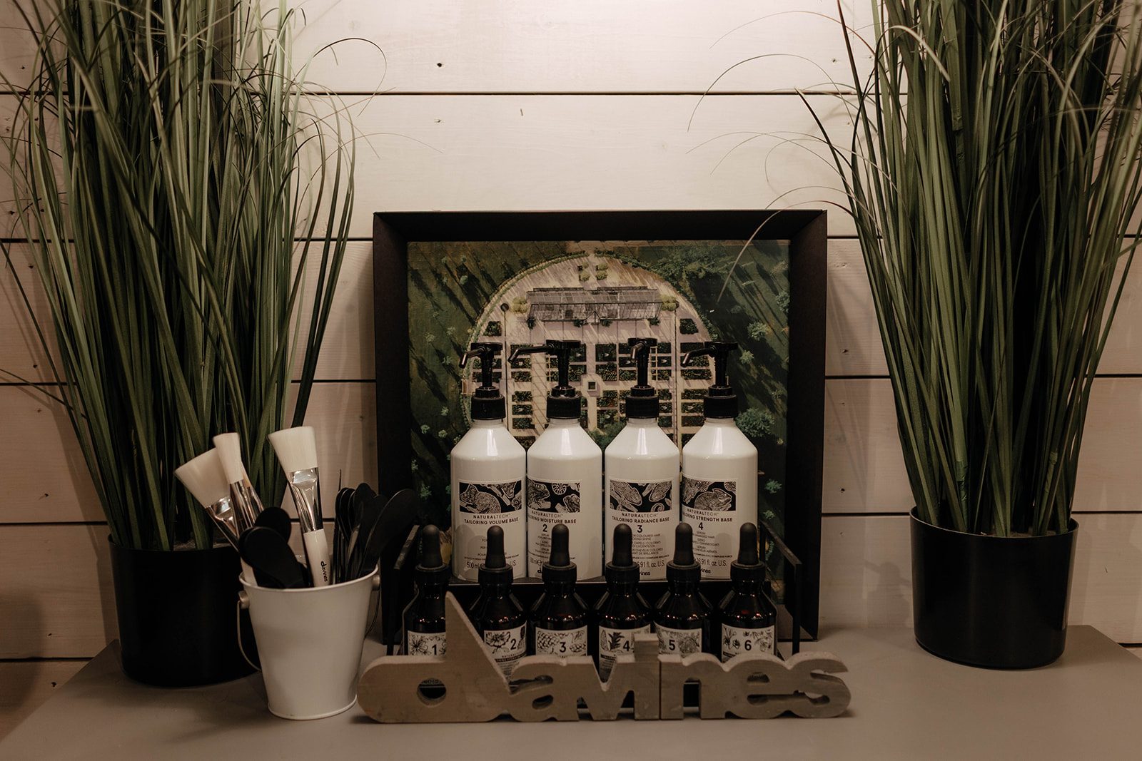 A Davines display on a grey shelf: white pump bottles, small dropper bottles, a bucket of colour brushes and a concrete Davines sign between two potted grasses