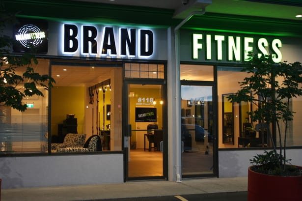 The lit BRAND and FITNESS signs over the glass storefront of unit #118 at dusk, with a yellow-walled studio visible through the window