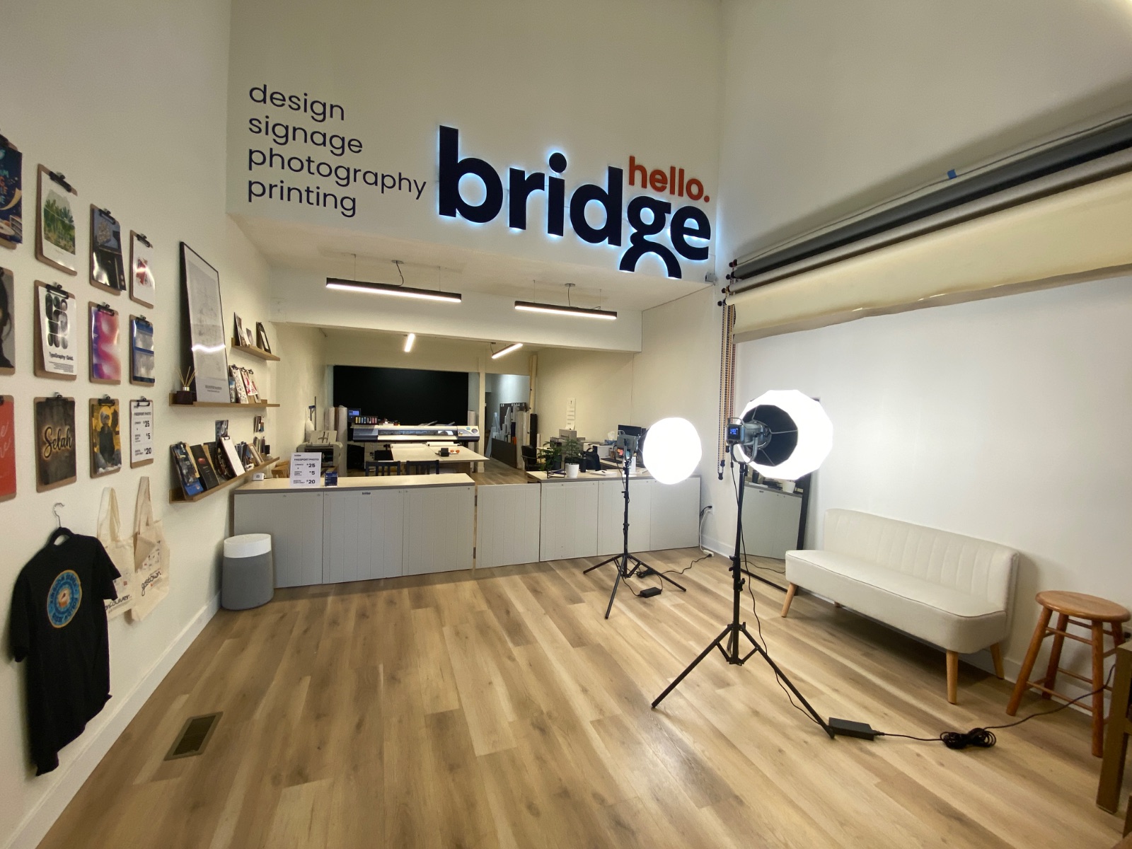 Inside Bridge Creative Studio: a bright room with a lit 'hello. bridge' sign, a photo backdrop and studio lights, a white sofa and samples of printed work on the walls