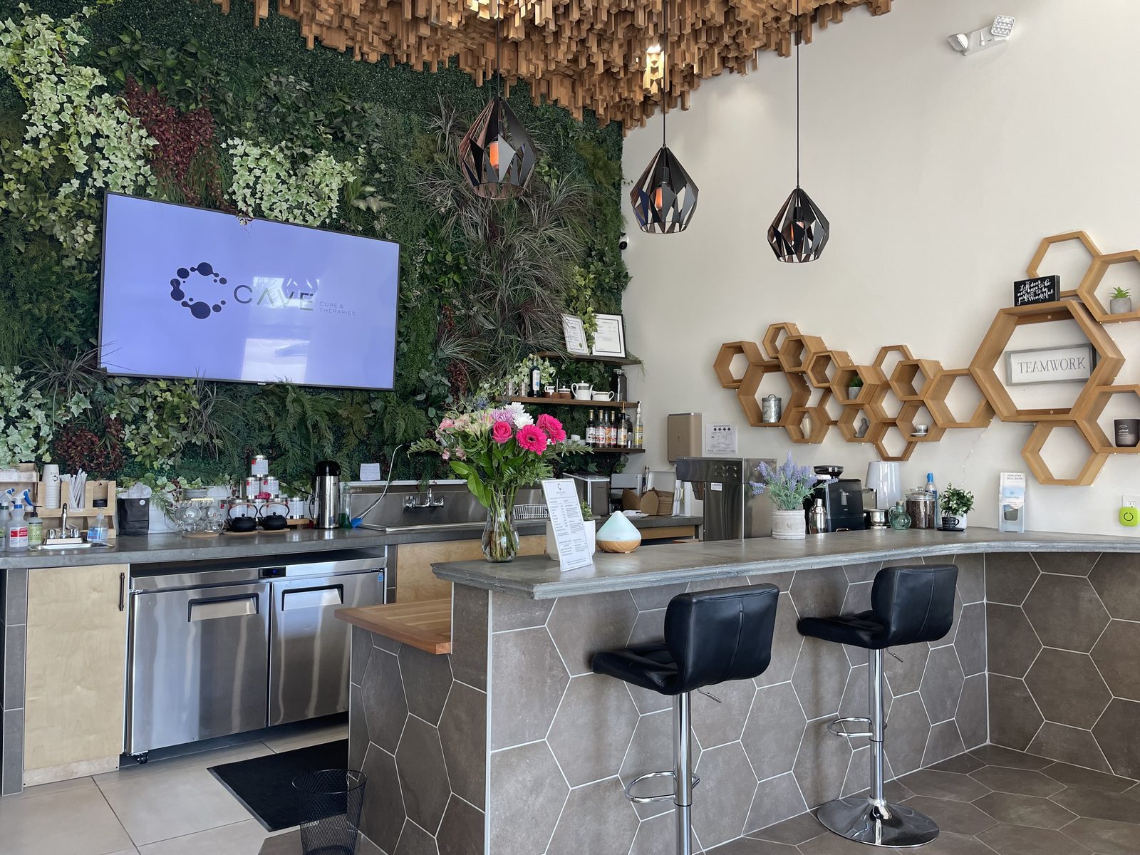 The front area at CAVE Cure & Therapies: a living plant wall with the CAVE logo on a screen, a café counter with tea tins and a coffee machine, hexagonal wooden shelves and two black bar stools