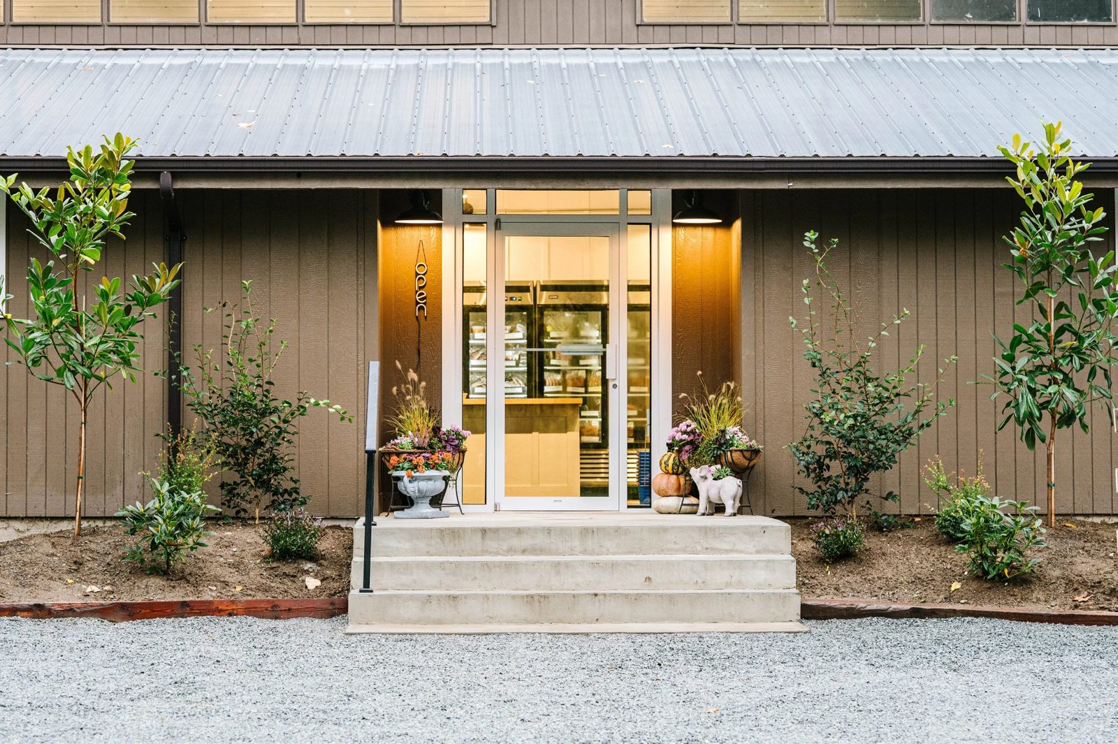 The Central Park Farms store: a brown-sided farm building with concrete steps up to a lit glass door, young trees and planters out front