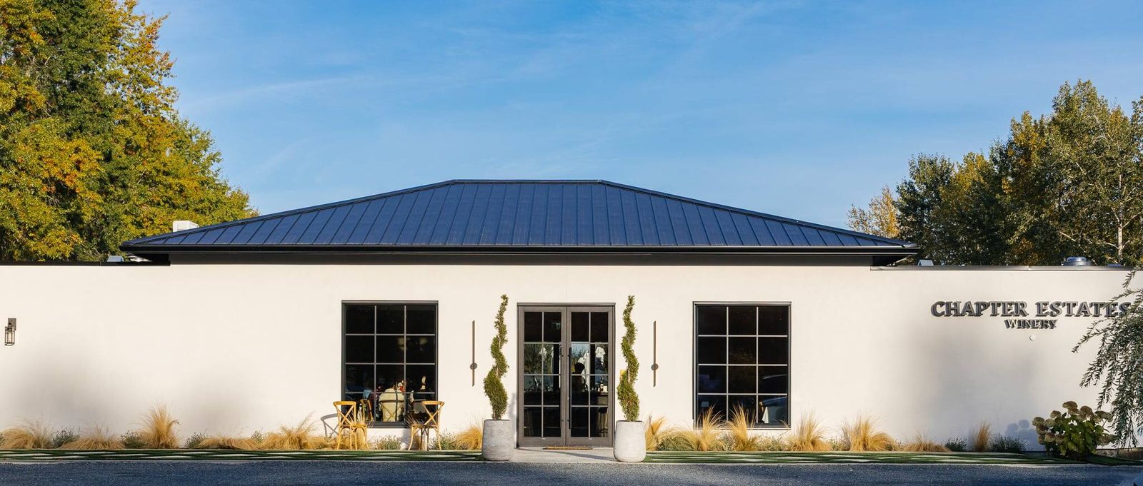 The white single-storey Chapter Estates Winery building with a dark standing-seam metal roof, double glass doors flanked by two spiral topiaries, and the winery name on the wall at right