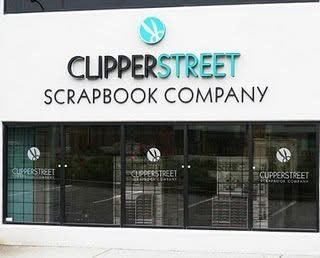 The Clipper Street Scrapbook Company storefront: a white wall with the shop's name in dark and teal lettering above a row of glass doors