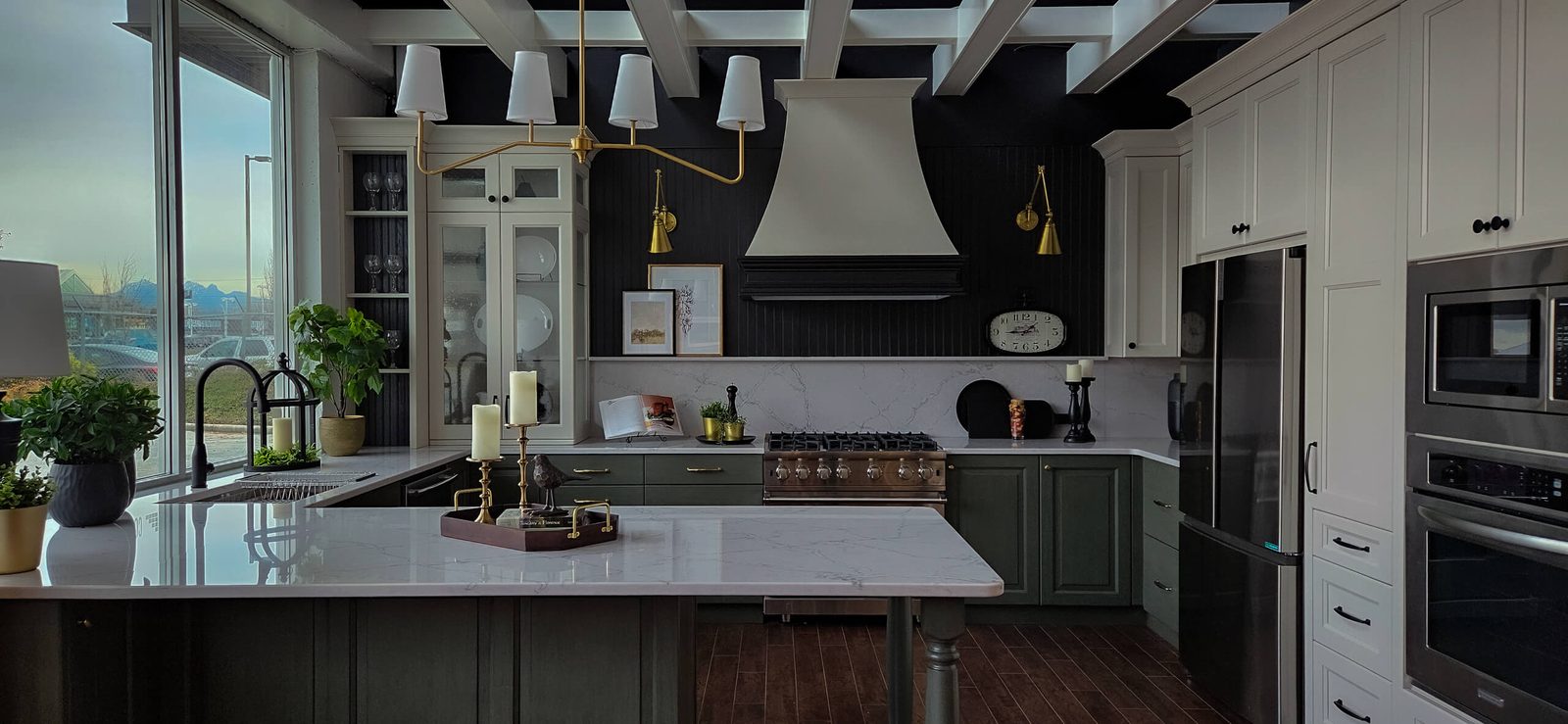 The Complete Cabinet Works showroom display kitchen: white upper cabinets, green-grey base cabinets, a black beadboard wall behind the range, a brass pendant light and a large window