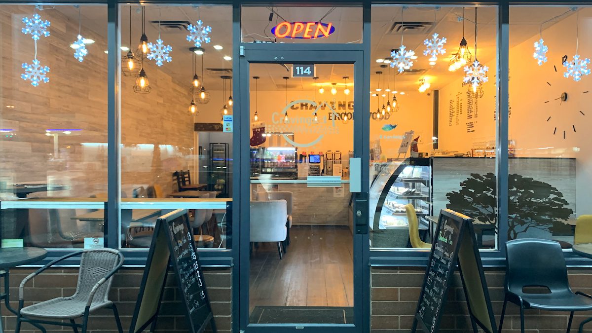 Cravings Coffee & Bakery's storefront at unit 114: glass front with an Open sign, window seating, a bakery case inside and patio chairs out front