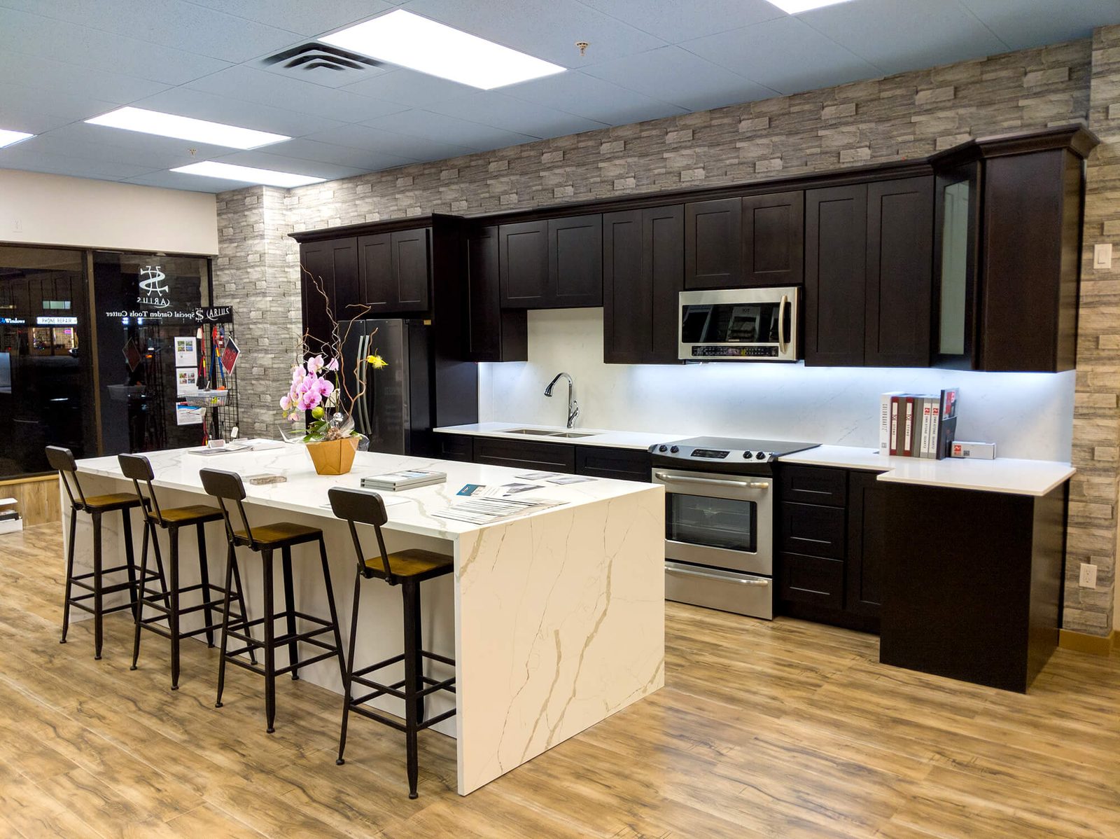 ECL Stonework's showroom: a display kitchen with a white waterfall-edge island and four bar stools, dark cabinets and a brick feature wall