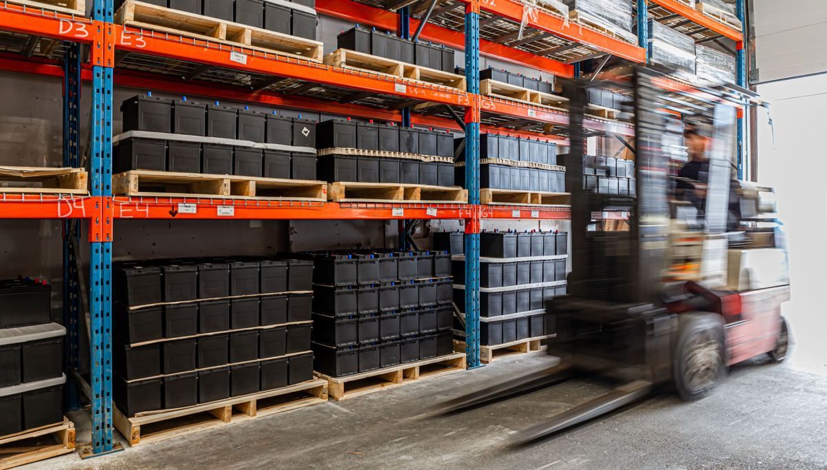 Pallets of boxed batteries stacked on orange warehouse shelving at Edmonds Batteries, with a forklift moving past