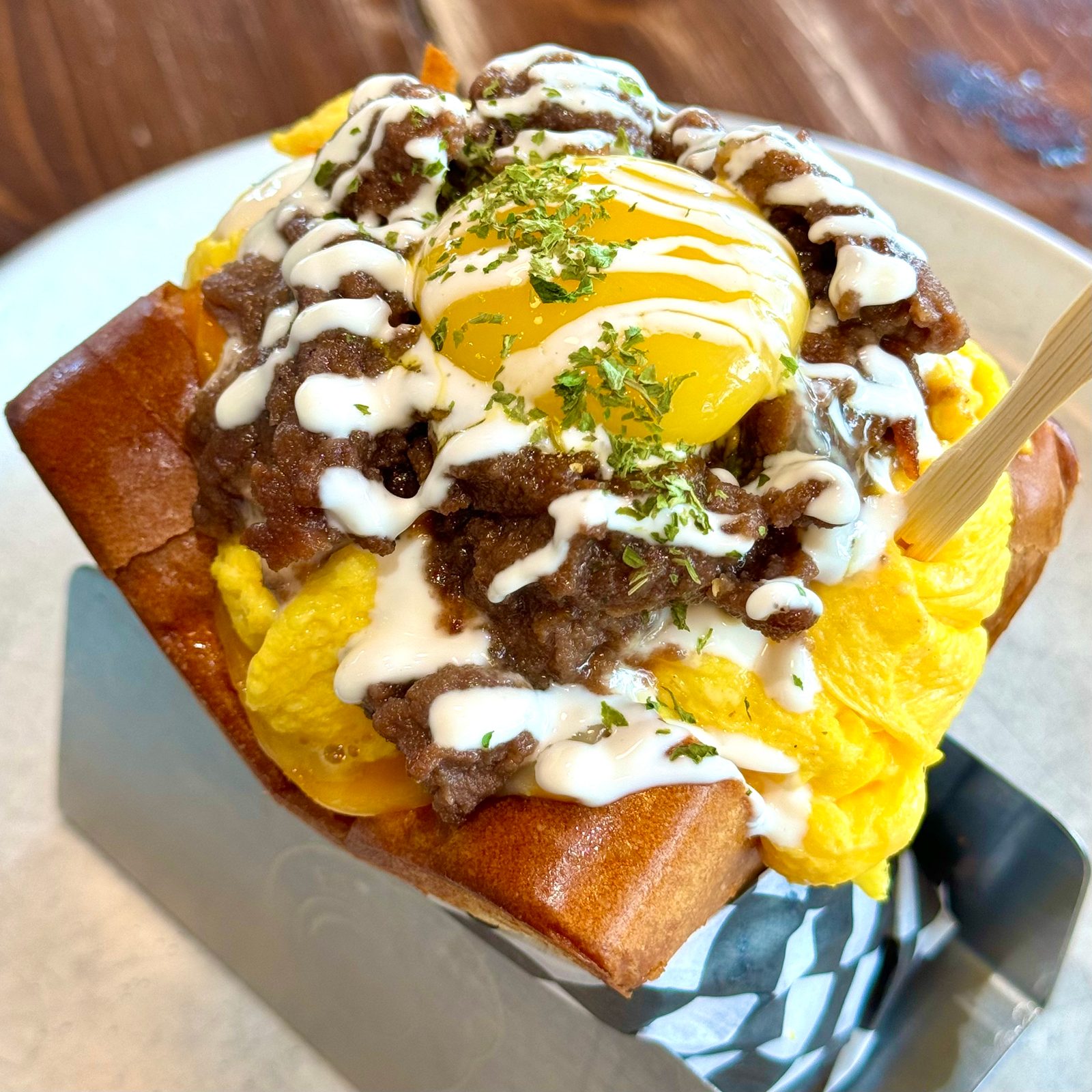 An Egg Bomb sandwich: a toasted bun piled with scrambled egg, beef, a soft yolk and squiggles of sauce, in a checkered paper-lined holder