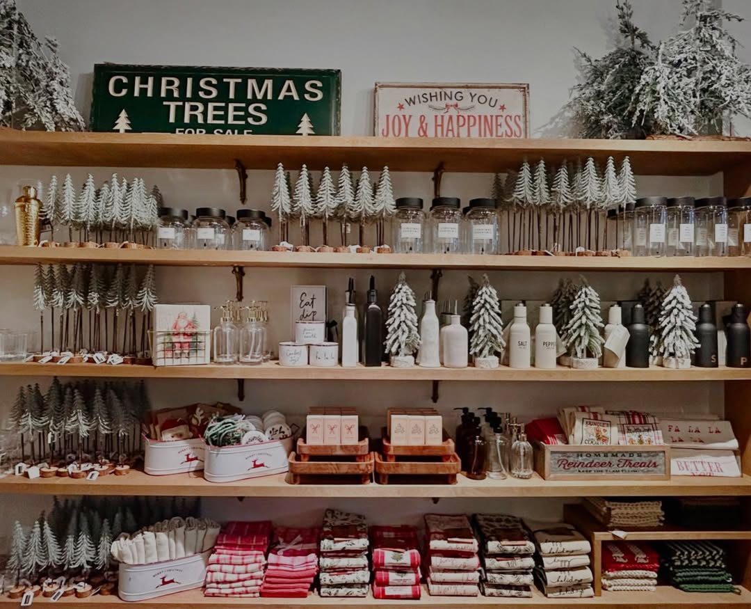 Shelves at Emilia & Rose Home Boutique lined with white bottlebrush trees, glass jars, candles and red-and-white tea towels under a Christmas Trees sign