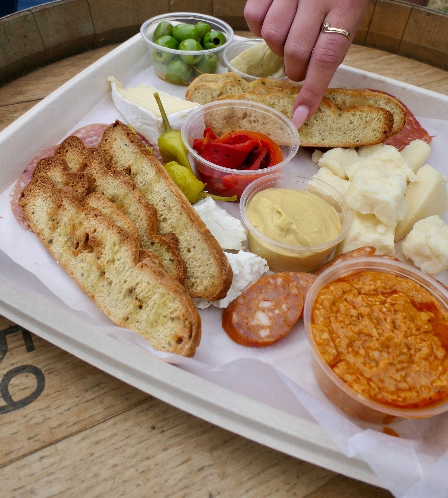 A picnic tray on a barrel top holding olives, brie, cured sausage, cheese curds, roasted red peppers, hummus, a red dip and toasted bread slices