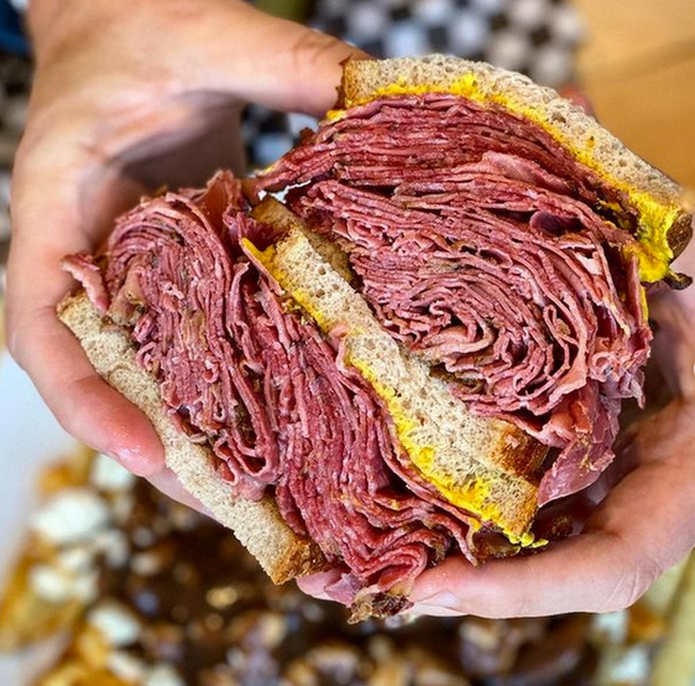 Two hands holding half an Estrella's Montreal smoked meat sandwich, the meat stacked thick on rye with mustard