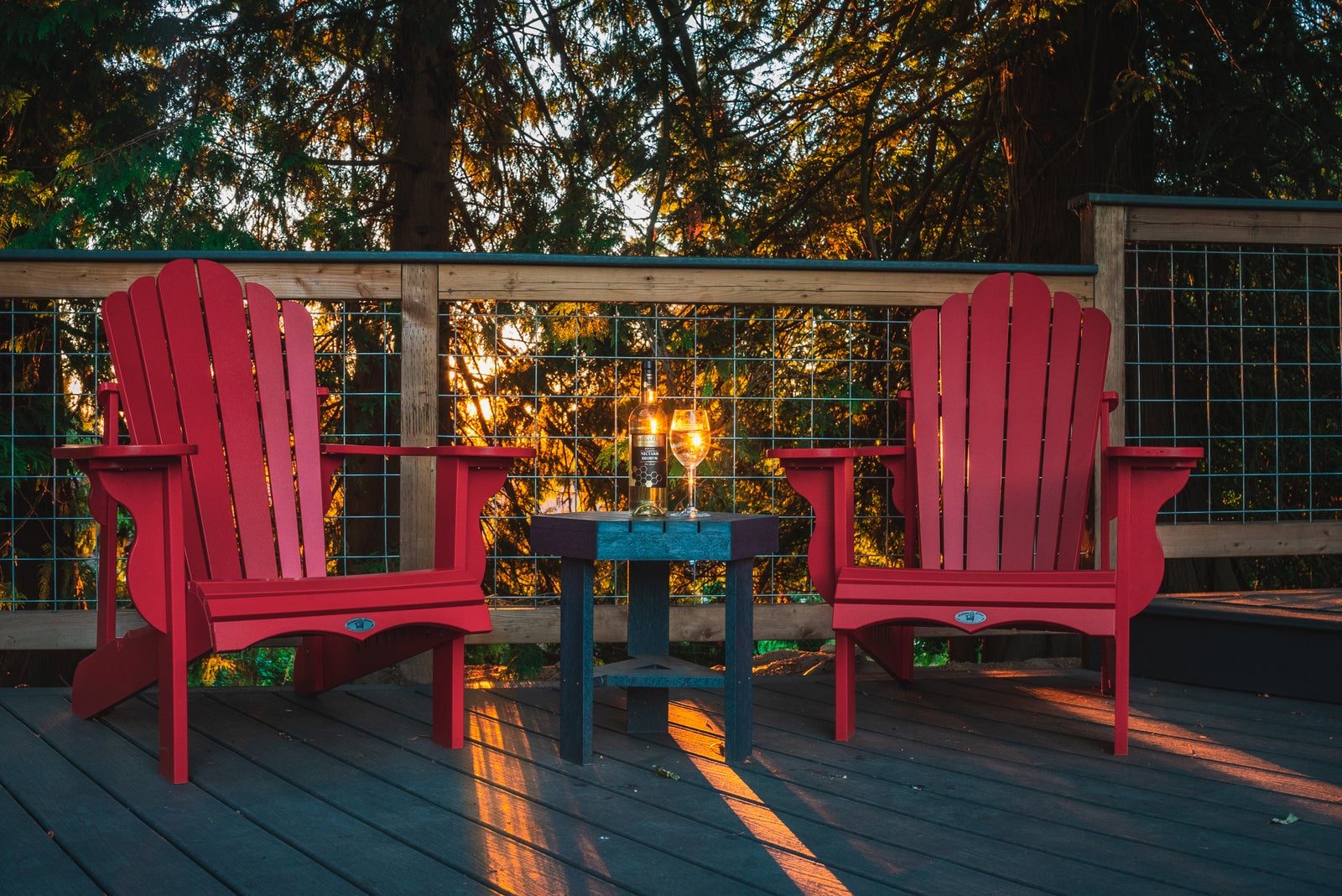 Two red Adirondack chairs on a wooden deck among tall trees at dusk, with a bottle and a glass of mead on the small table between them