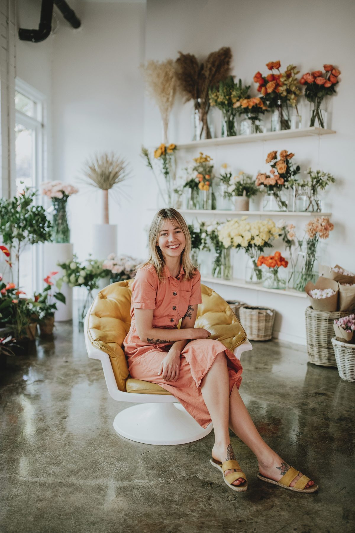 Floralista's owner sitting in a yellow chair inside the bright white flower studio, with shelves of vases and buckets of fresh flowers behind her