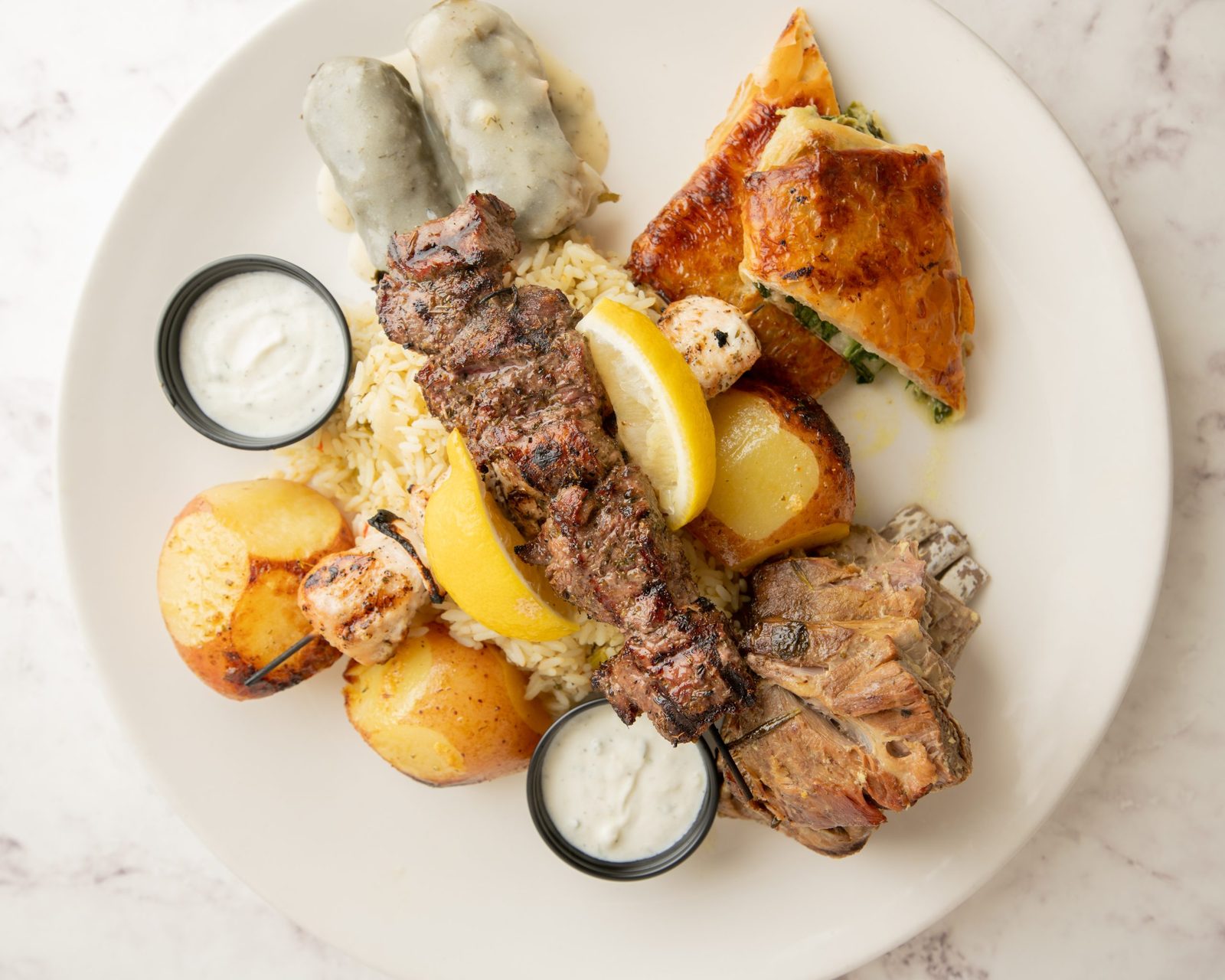 A Galini plate with a lamb souvlaki skewer, spanakopita, dolmades, roast potatoes and tzatziki