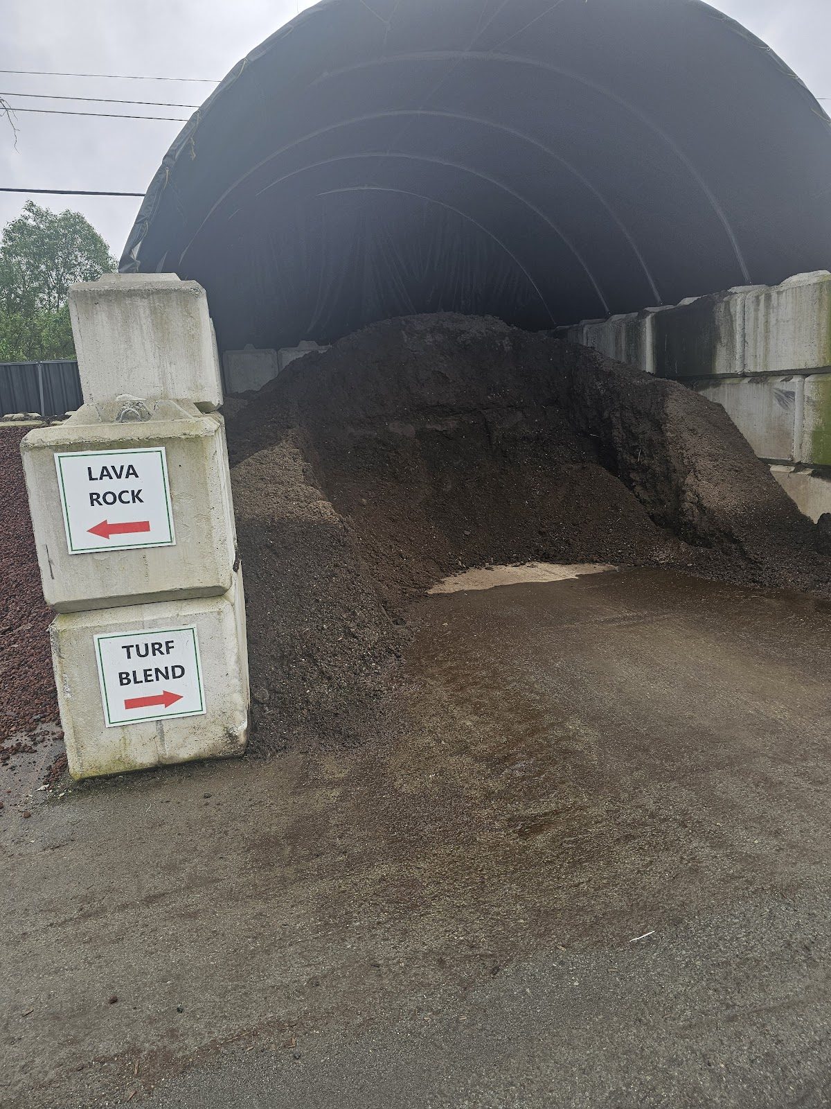 A big pile of dark soil under a covered bay at Green Valley's yard, with concrete-block signs pointing to Lava Rock and Turf Blend