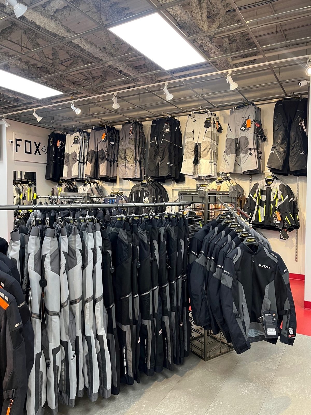Racks of motorcycle riding jackets and pants in grey, black and hi-vis in Holeshot's apparel department, with more gear hung on the walls