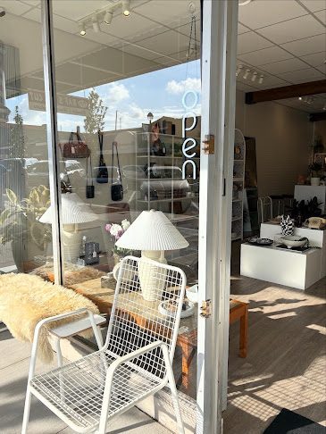 Looking through Homebody Goods' open front door on Fraser Highway: a white wire chair, a lamp and shelves of home goods in a bright shop