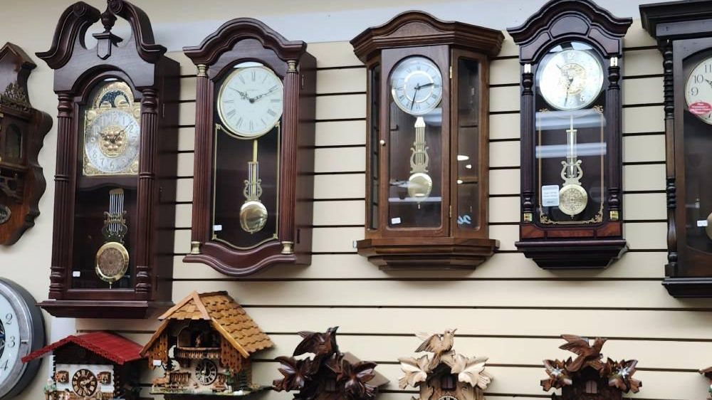 A wall of wooden pendulum clocks with brass dials in the It's About Time & Jewelry showroom, with cuckoo clocks hanging in a row below them