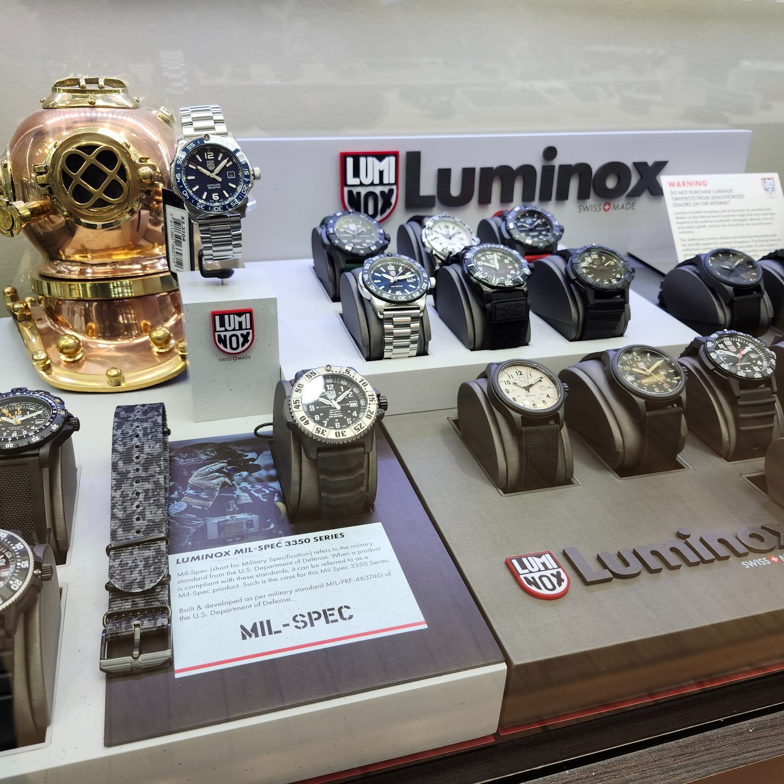 A glass display case of Luminox dive and field watches on grey stands, with a polished copper and brass diving helmet at one end