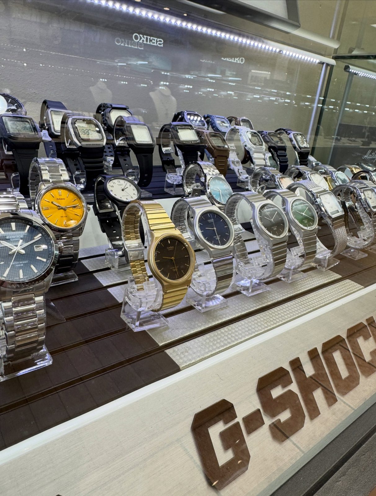 Rows of Casio and Edifice watches on clear stands inside a lit display case with a G-Shock sign along the bottom