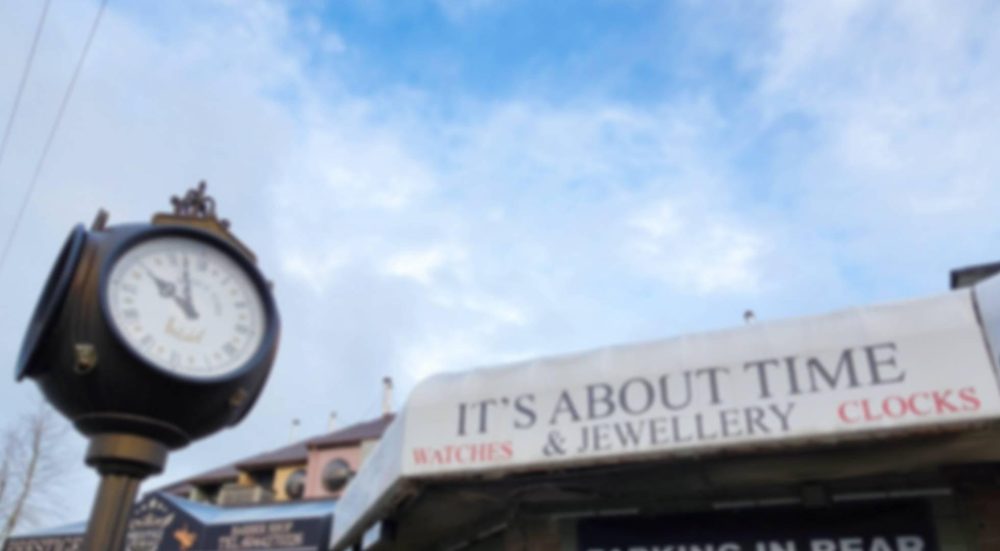 The shop's white awning reading It's About Time & Jewellery, Watches, Clocks, with a black and gold street clock on a pole beside it
