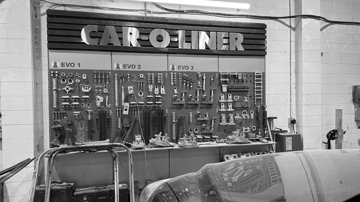 A black-and-white photo inside Jan's Precision Auto Body: a wall of frame-measuring and pulling fixtures under a Car-O-Liner sign, with a car hood in the foreground