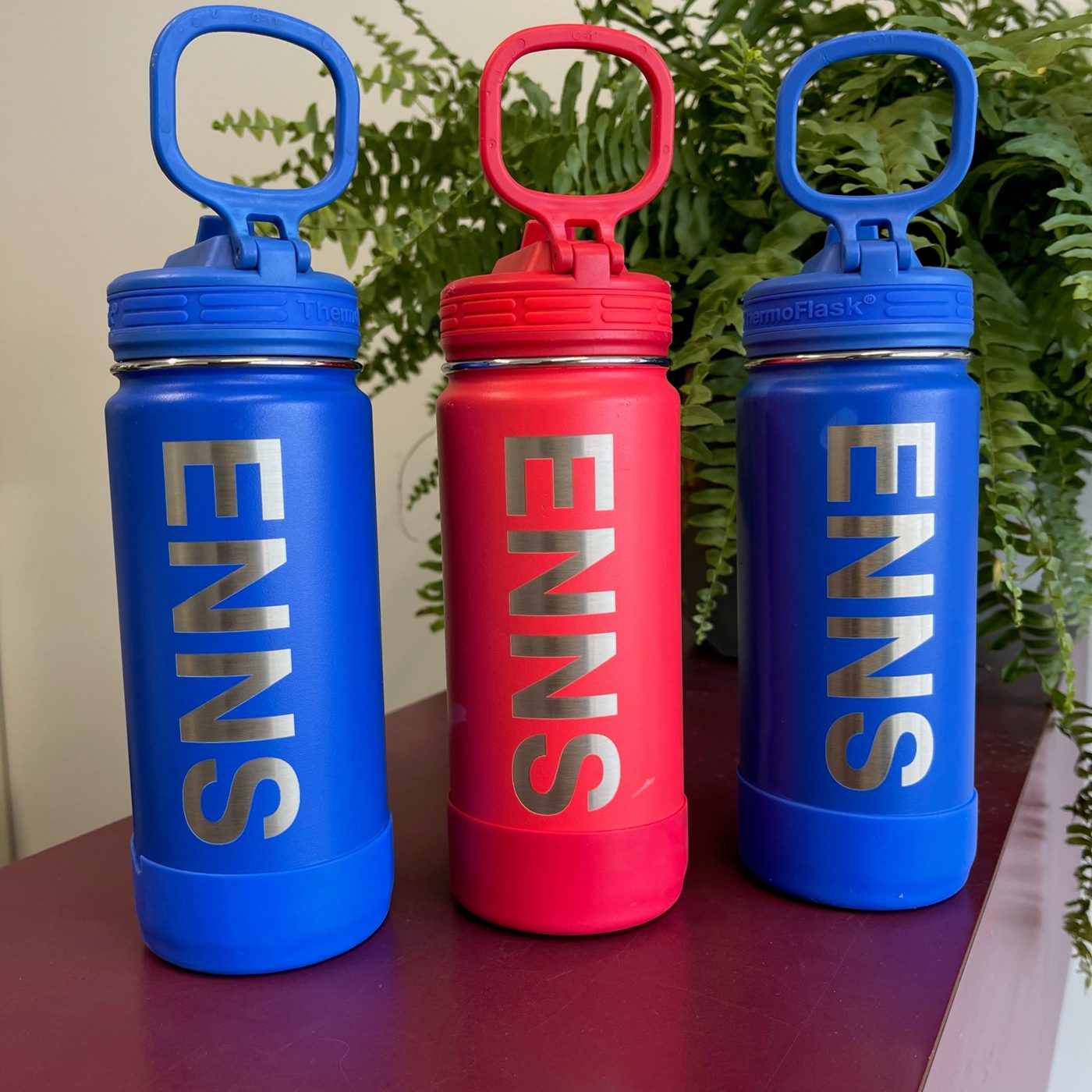 Three kids' water bottles, two blue and one red, each laser-engraved with a family name, from JMB Engraving