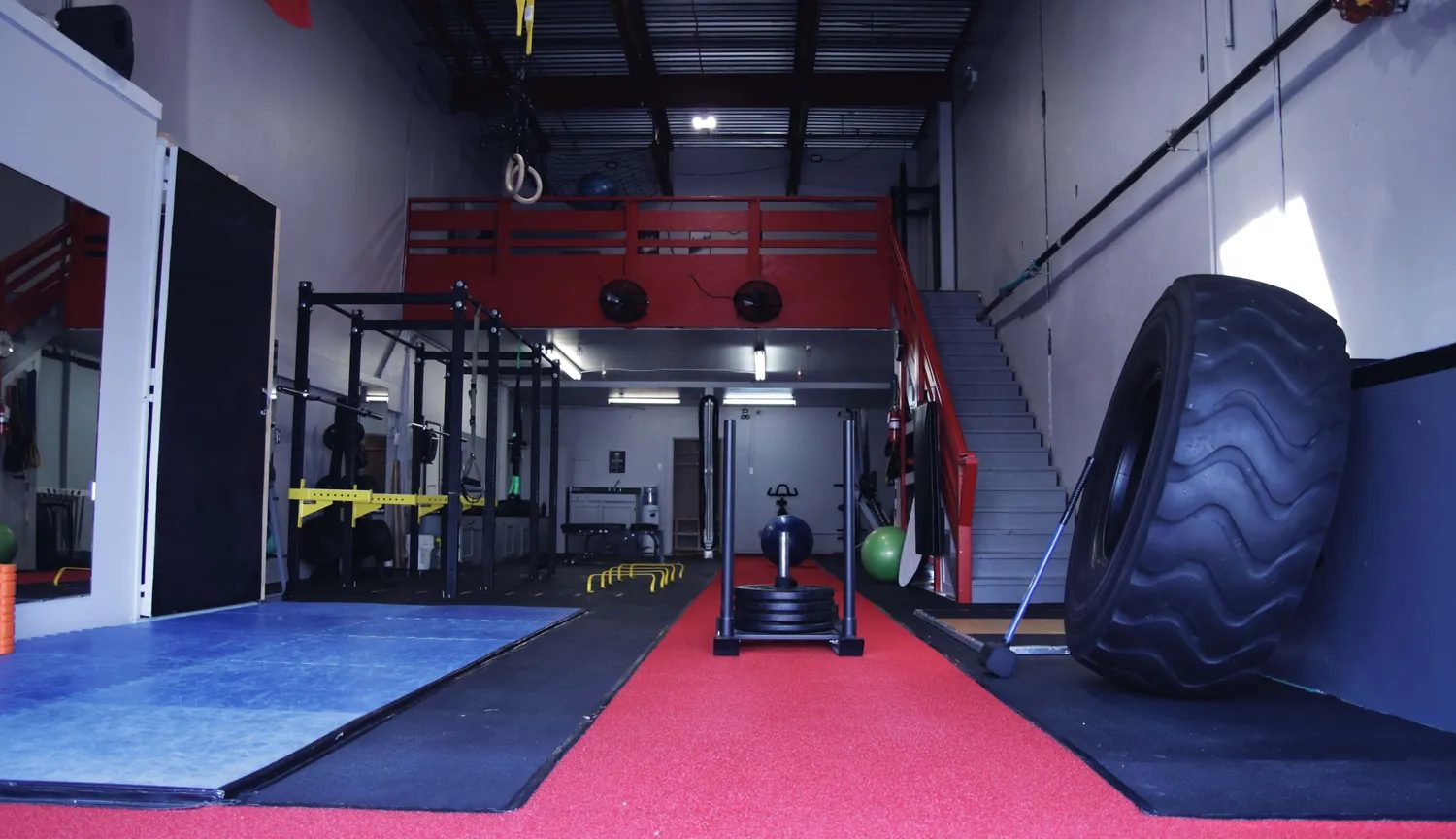 The main training floor at Kenaz Training: a red turf sprint lane with a push sled, power racks on the left, a large tire on the right and a red-railed loft at the back