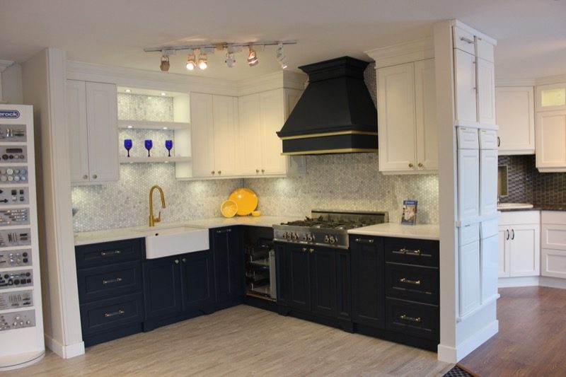 A display kitchen in the Kitchen Concepts showroom: navy lower cabinets with brass pulls, white upper cabinets, a farmhouse sink with a brass tap and a black range hood banded in brass