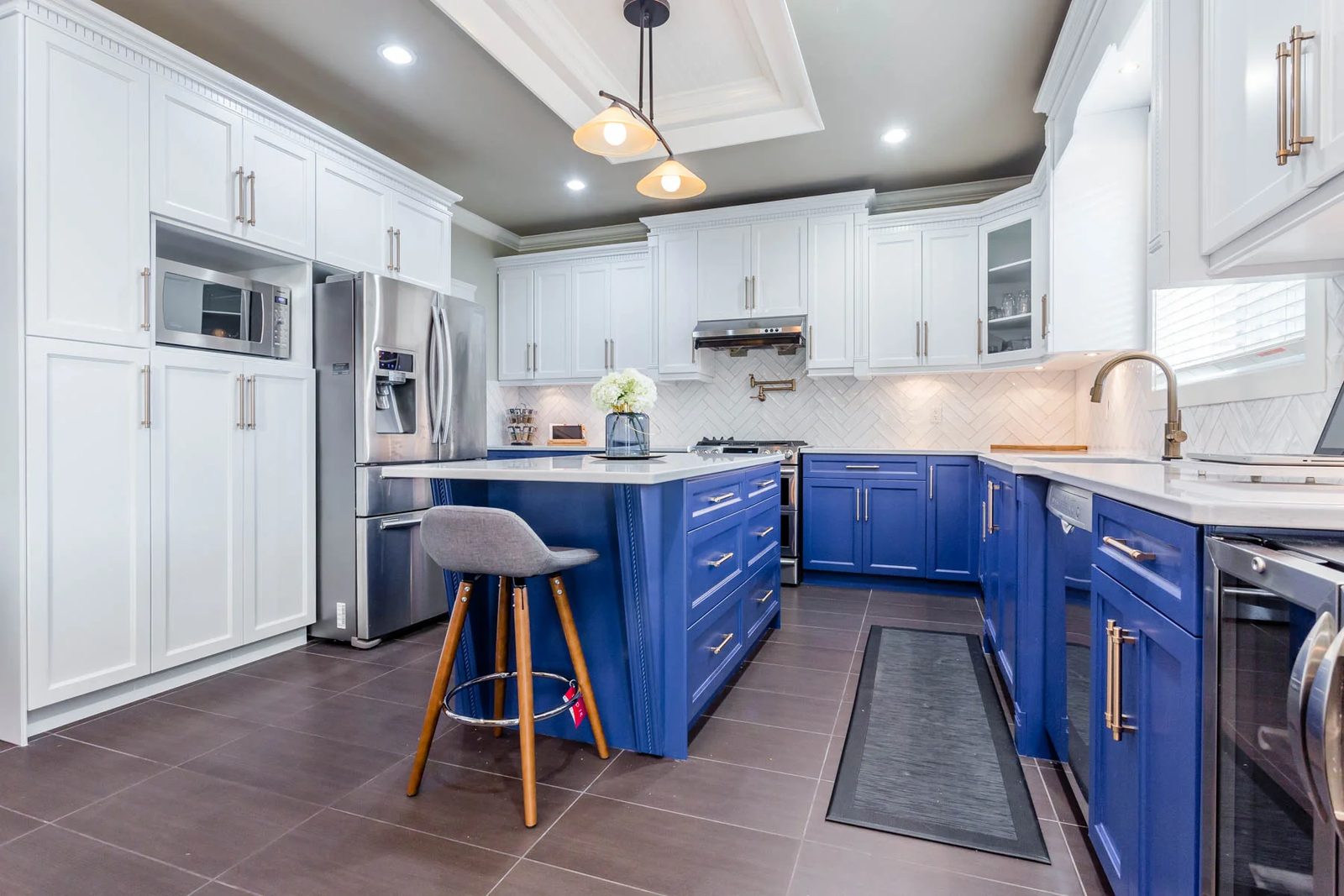 A kitchen with white upper cabinets and a deep blue island and lower cabinets, brass pulls, a herringbone tile backsplash and dark floor tiles
