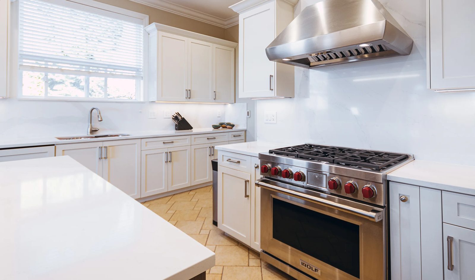 A bright kitchen with white shaker cabinets, white quartz counters, a large stainless hood and a six-burner range with red knobs