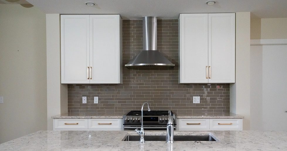 Two white cabinets with gold pulls on either side of a stainless range hood over a grey glass-tile backsplash