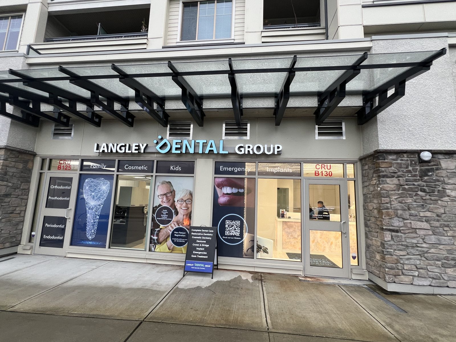 The street-level front of Langley Dental Group, unit B130: a lit Langley Dental Group sign over glass windows with service posters, a sandwich board and a stone-faced wall beside the door