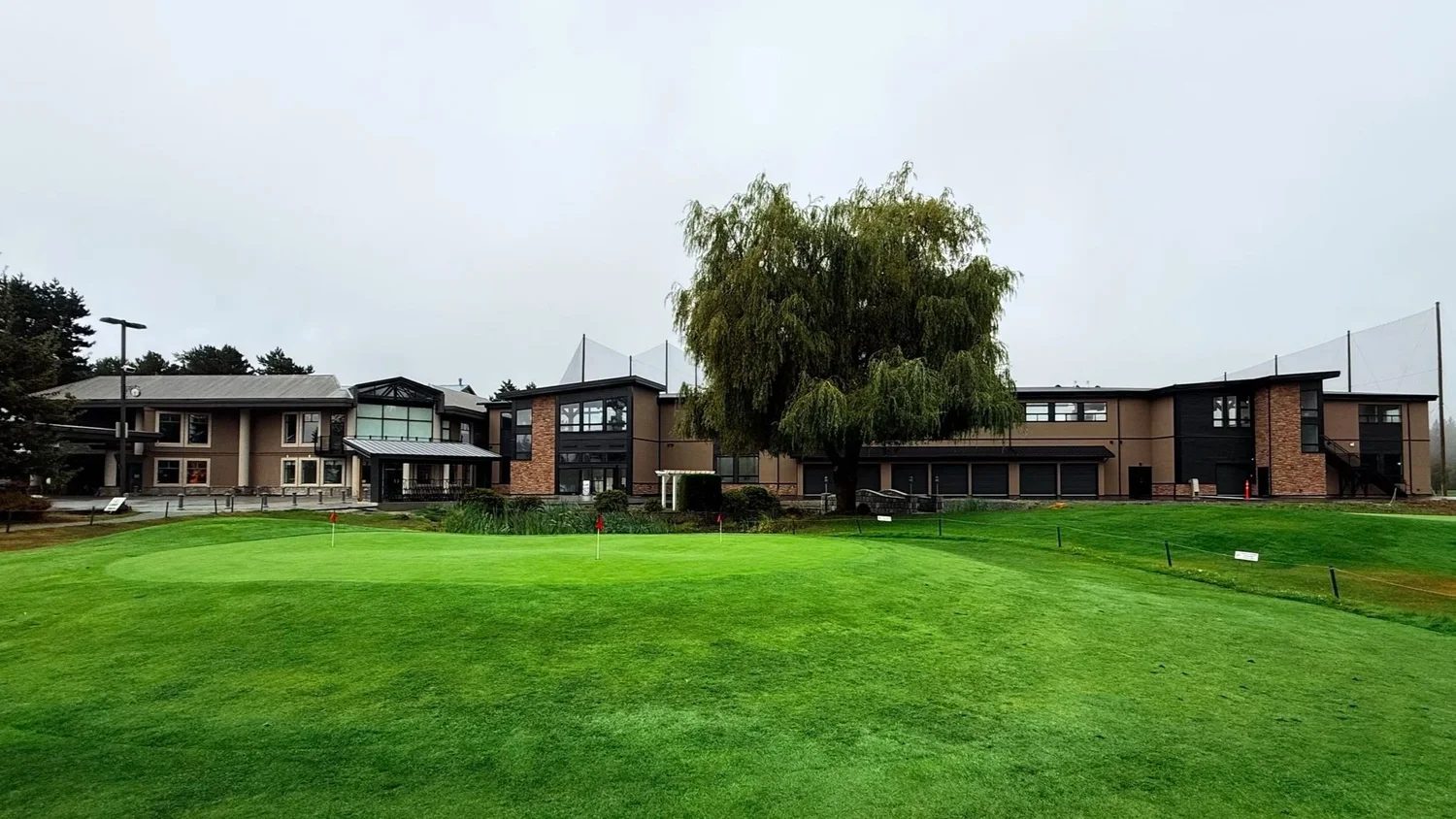 The Langley Golf Centre clubhouse and driving range building behind a practice green with red flags, under a big weeping willow on an overcast day
